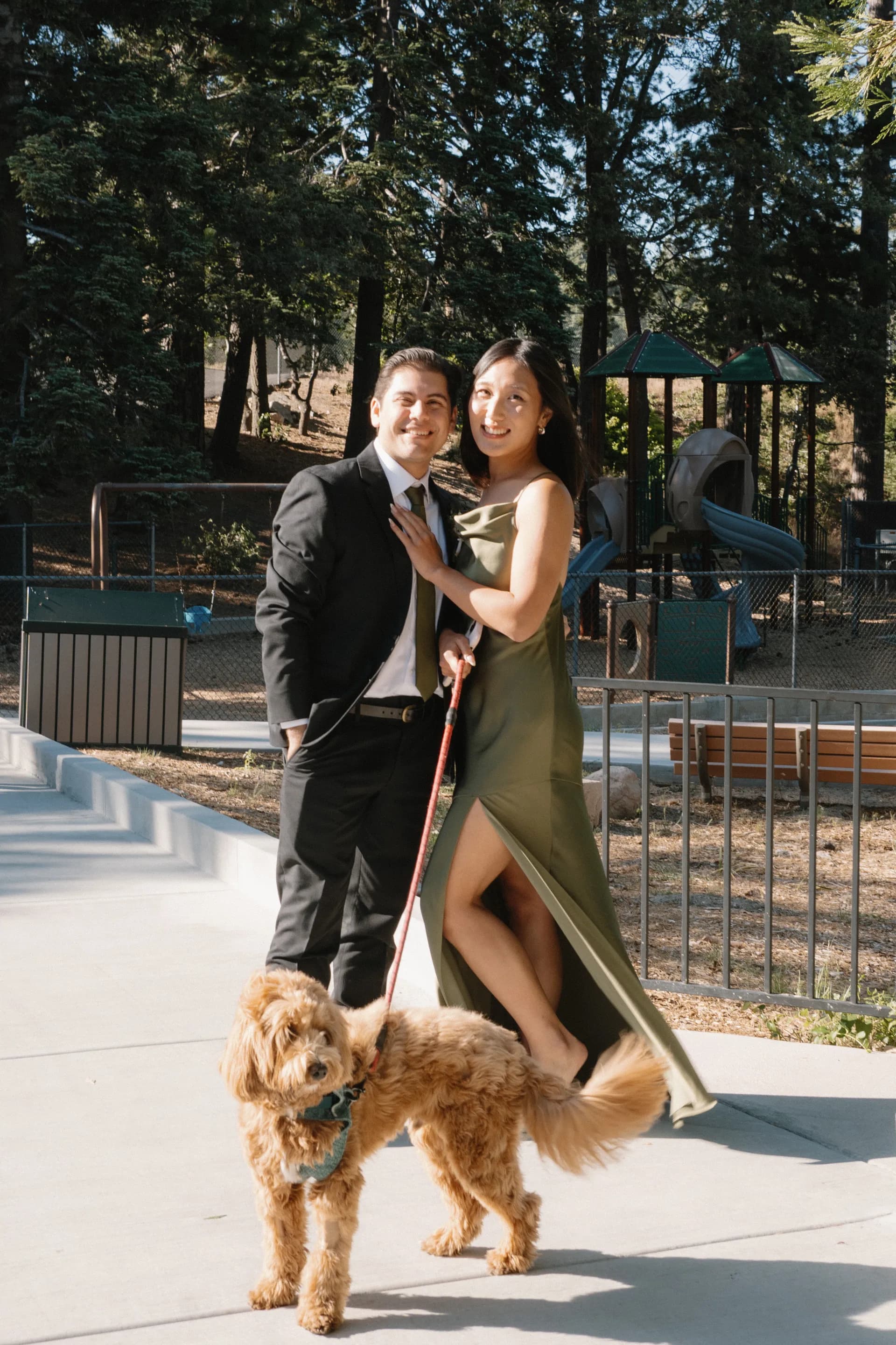 JP and Rachel dressed up and smiling in a park, with Charlie the goldendoodle on a red leash in front of them